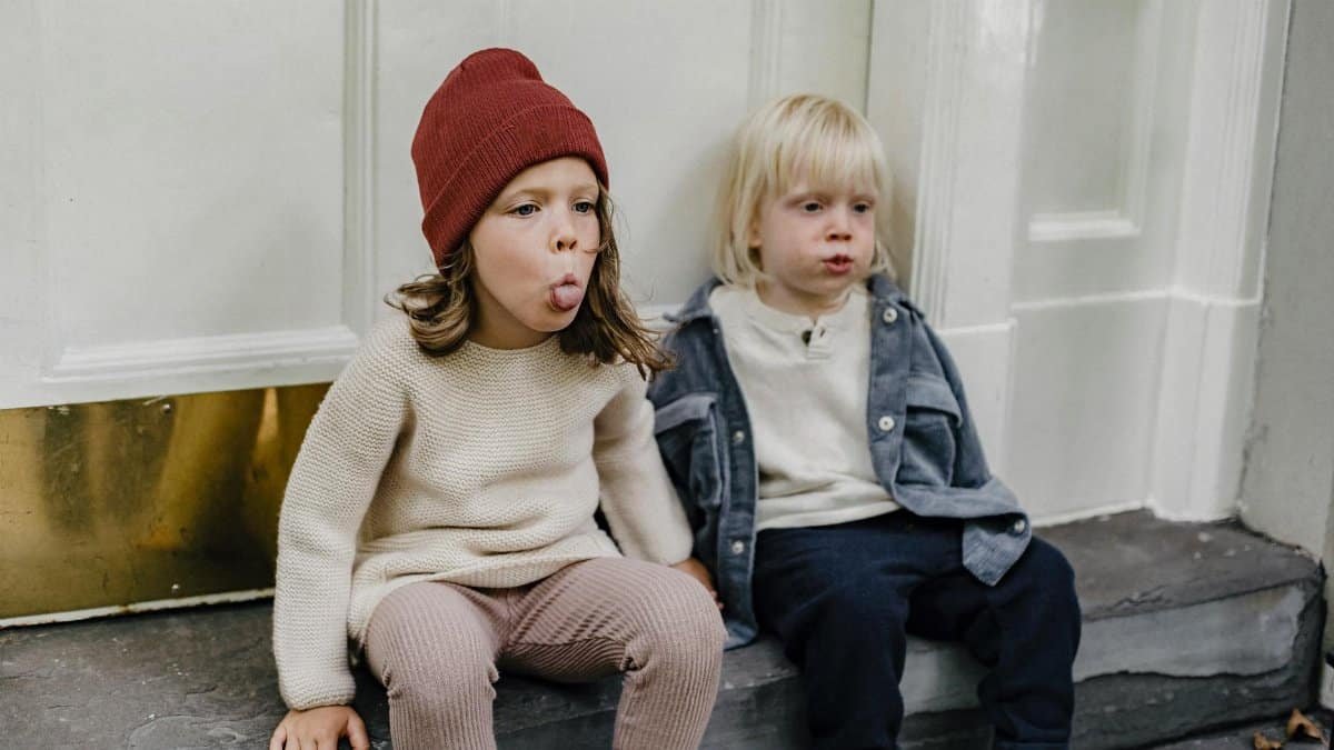 Adorable girl in hat showing tongue while sitting on step with cute little boy near entrance of building on street