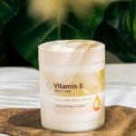 Vitamin E skin care cream jar displayed on a natural stone surface surrounded by lush green leaves.