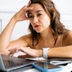 A woman with a frustrated expression, hand on head, working on a laptop.