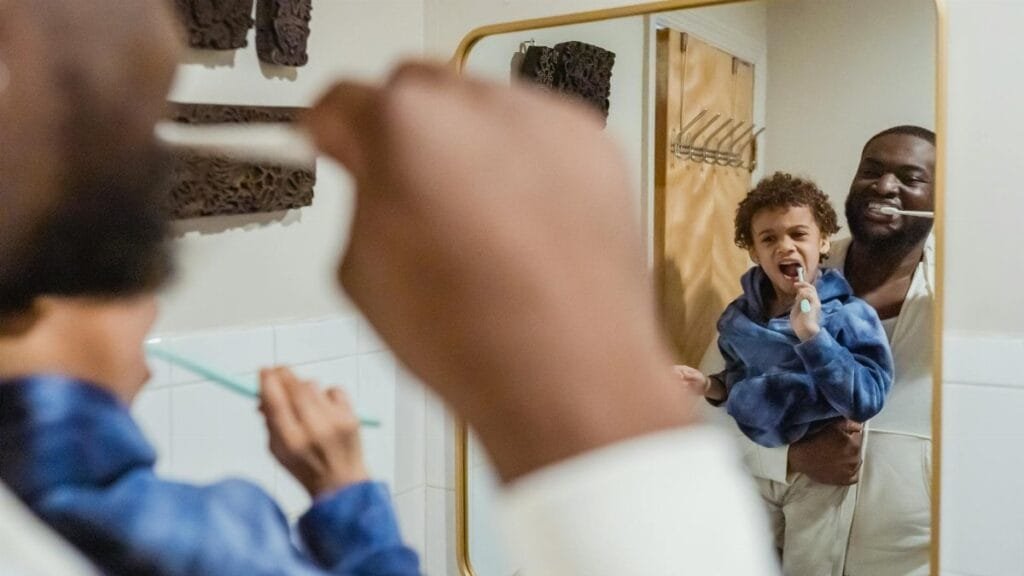Positive African American male carrying cute son on hand and brushing teeth together against mirror in bathroom