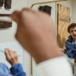 Positive African American male carrying cute son on hand and brushing teeth together against mirror in bathroom