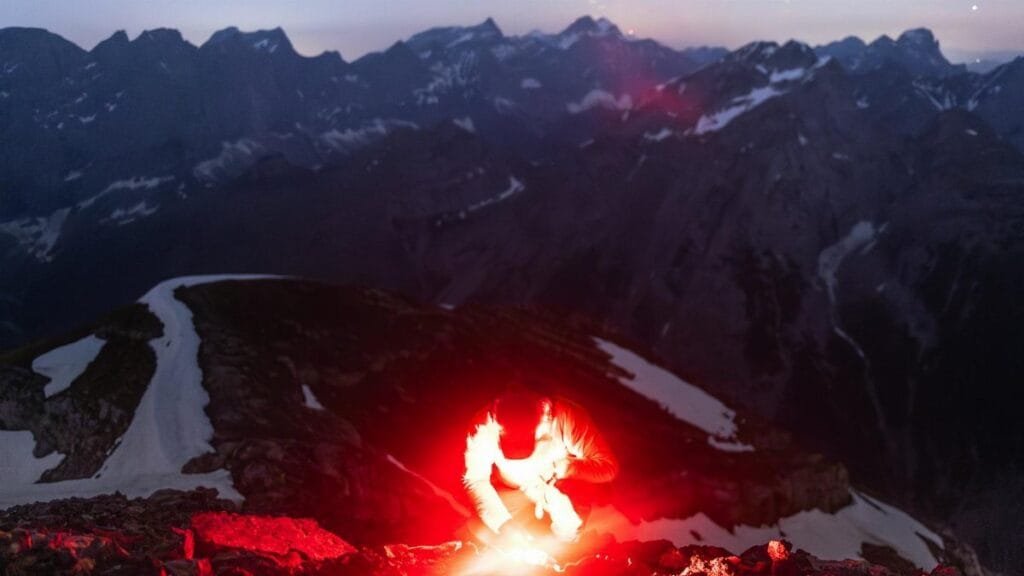 Hiker lit by flare in the mountainous terrain under a starry sky.