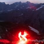 Hiker lit by flare in the mountainous terrain under a starry sky.