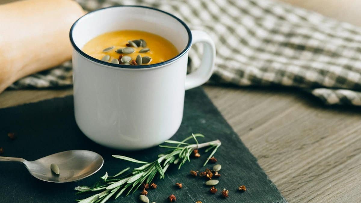 Warm pumpkin soup garnished with seeds and herbs in an enamel mug, perfect for fall comfort food.