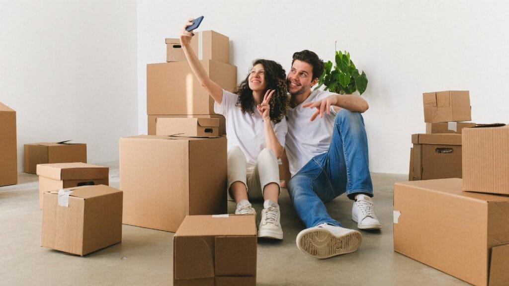 Happy couple sitting among moving boxes taking a selfie inside their new home.