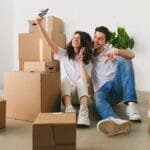Happy couple sitting among moving boxes taking a selfie inside their new home.