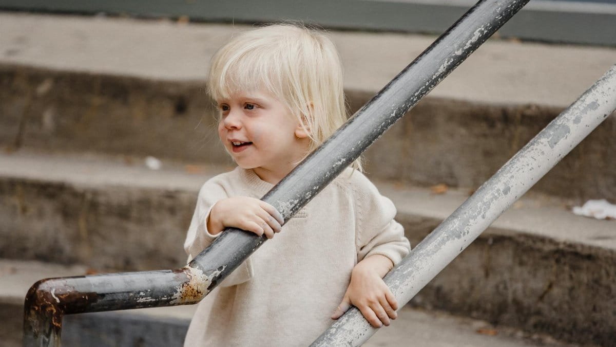 Side view of cheerful adorable little girl in casual outfit standing on staircase near railing and looking away