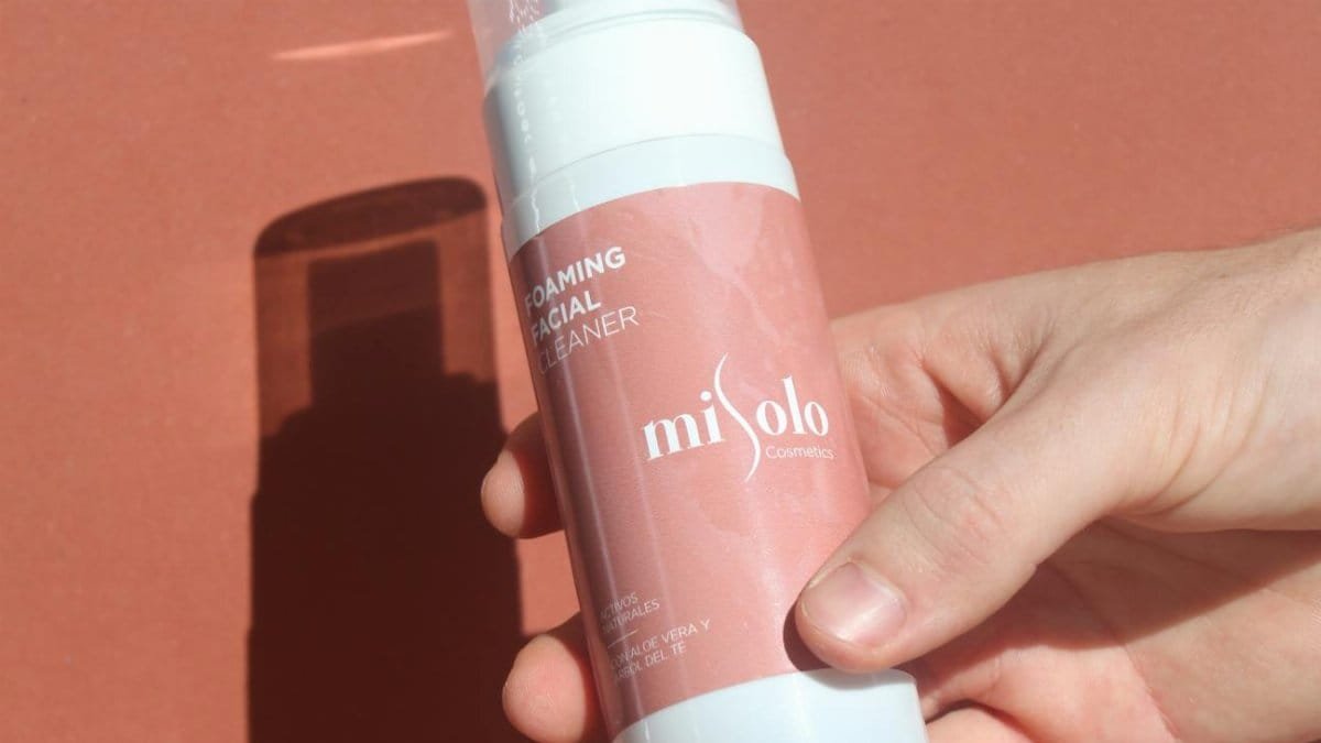 A hand holding a foaming facial cleanser bottle by Misolo Cosmetics against a soft pink background.