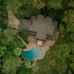 A scenic aerial shot of a house surrounded by lush forest and a swimming pool.