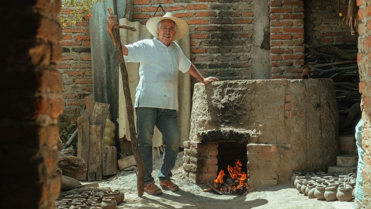 Elderly artisan in a straw hat managing a traditional fire clay oven in a rustic pottery workshop.