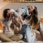 Joyful parents playing with their child on a bed in a warmly lit bedroom.