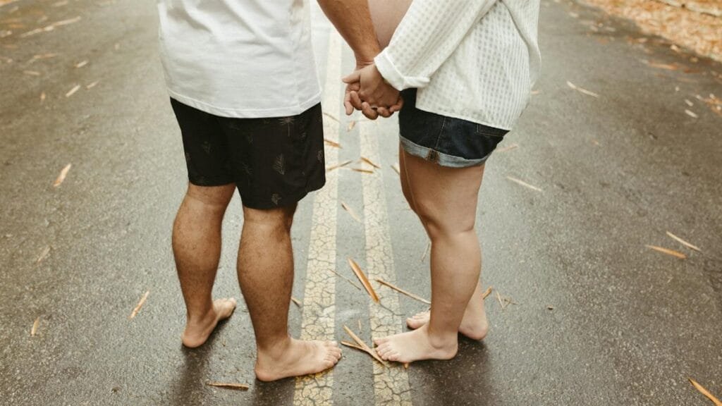 Barefoot expecting couple holding hands on a leaf-strewn road, symbolizing unity and anticipation.