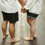 Barefoot expecting couple holding hands on a leaf-strewn road, symbolizing unity and anticipation.