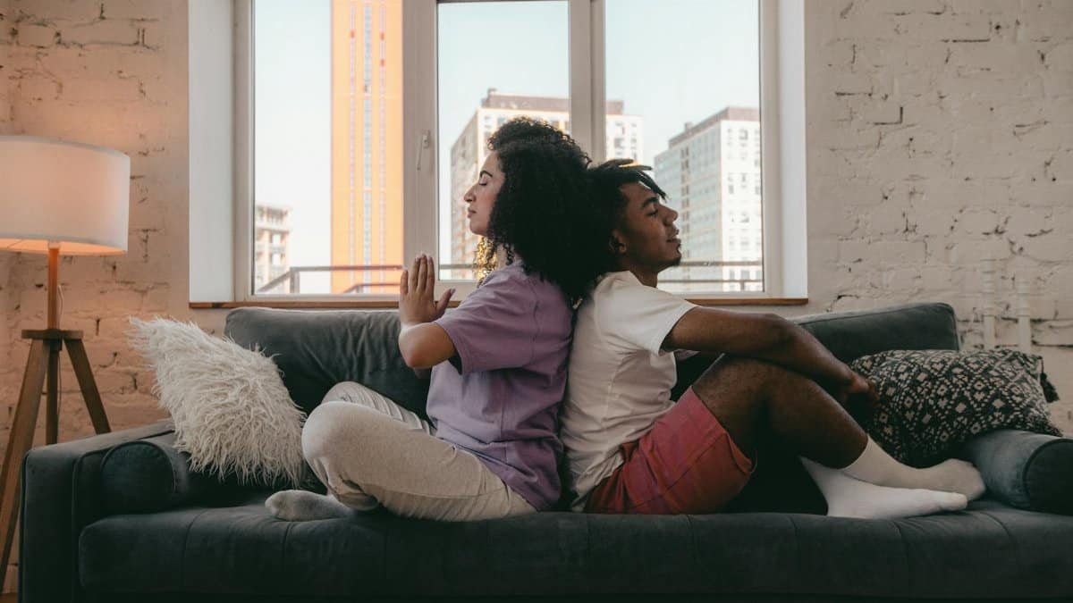 Interracial couple practicing meditation back-to-back on a couch. Calm and peaceful ambiance.
