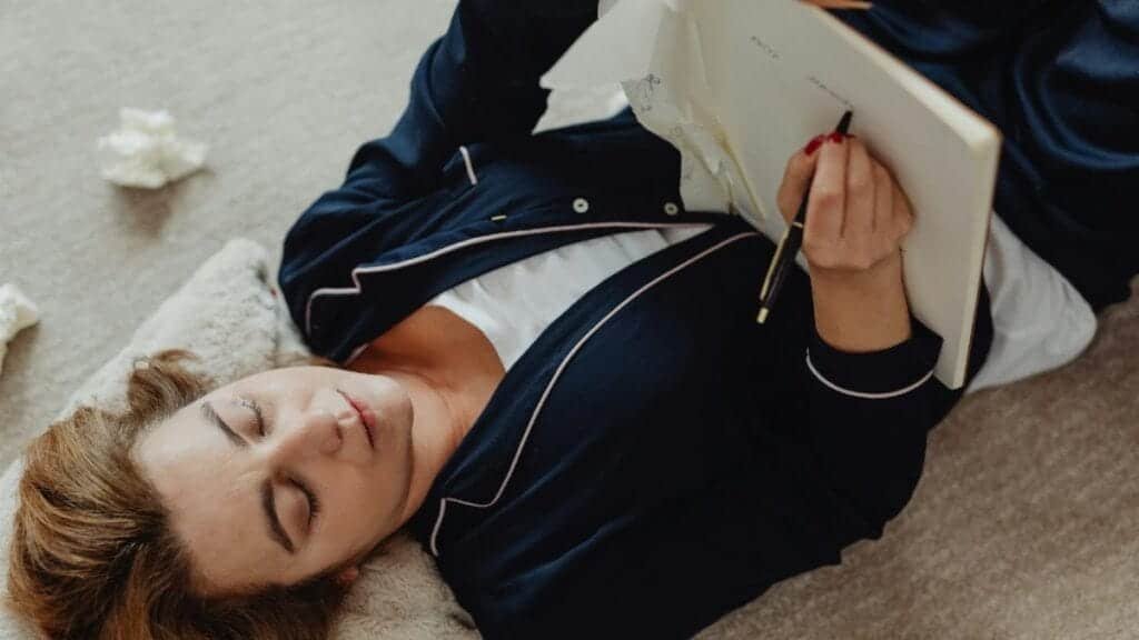 An adult woman relaxes while writing in a notebook with crumpled papers around her.