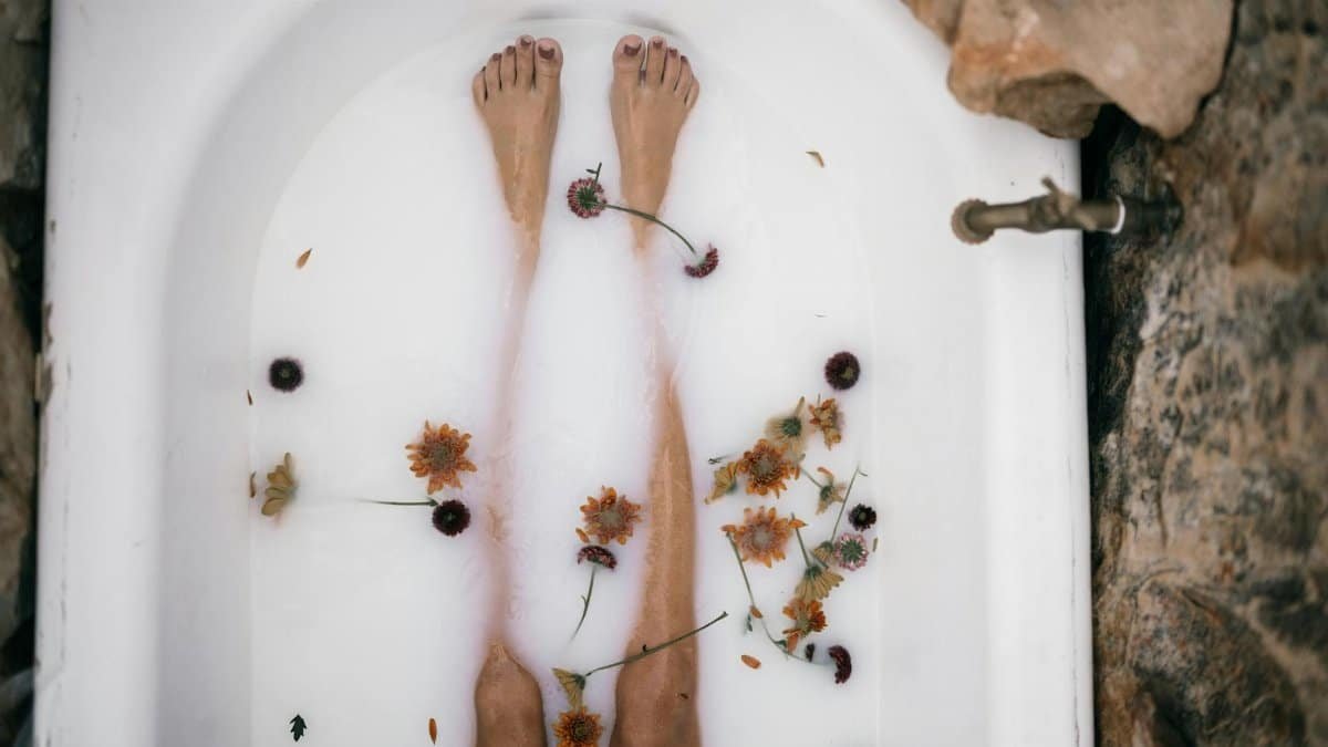 Top view of a woman's bare legs in a bathtub filled with milk and scattered flowers for a soothing bath experience.