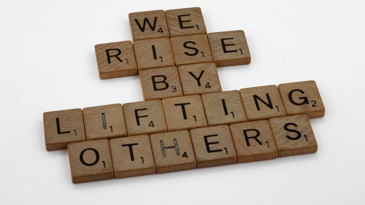 Scrabble tiles forming the quote 'We rise by lifting others' on a white background.