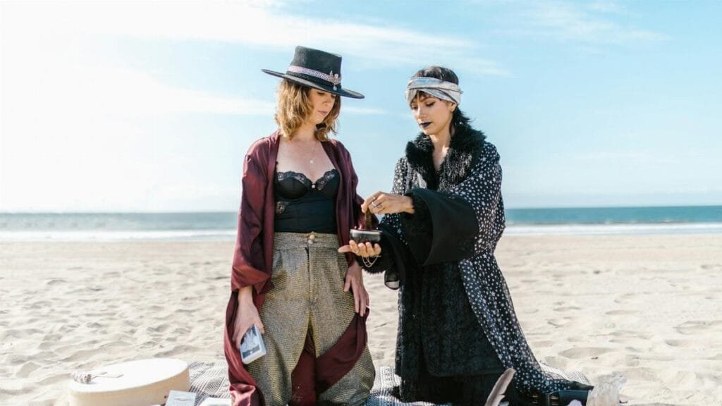 Two women engage in a spiritual ritual on a sunny beach, surrounded by crystals.