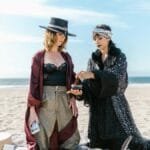 Two women engage in a spiritual ritual on a sunny beach, surrounded by crystals.