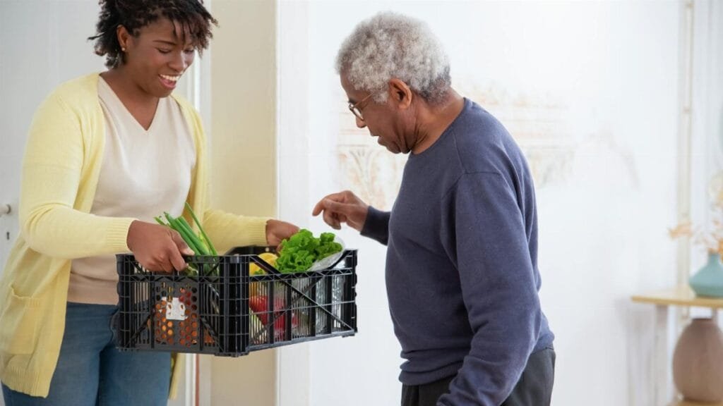 A young adult woman helps a senior man by carrying a crate of fresh vegetables indoors, symbolizing care and support.