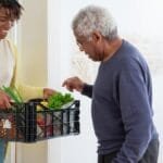 A young adult woman helps a senior man by carrying a crate of fresh vegetables indoors, symbolizing care and support.
