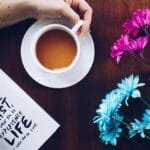 A cozy setup with tea, flowers, and an inspirational quote book on a wooden table.