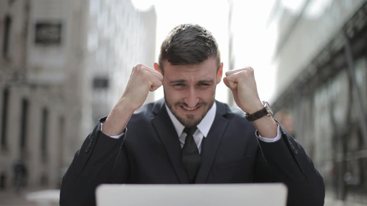 A joyful businessman in a suit triumphantly celebrates success on his laptop outdoors.