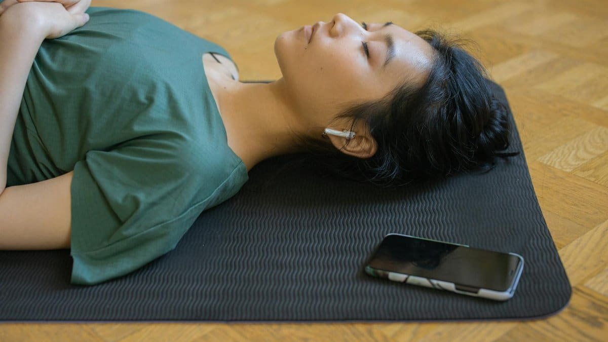 Asian woman resting on yoga mat with wireless earbuds and smartphone indoors.