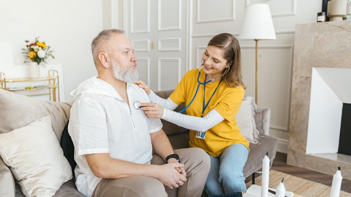 A nurse uses a stethoscope for a home check-up on a senior adult in a cozy living room.
