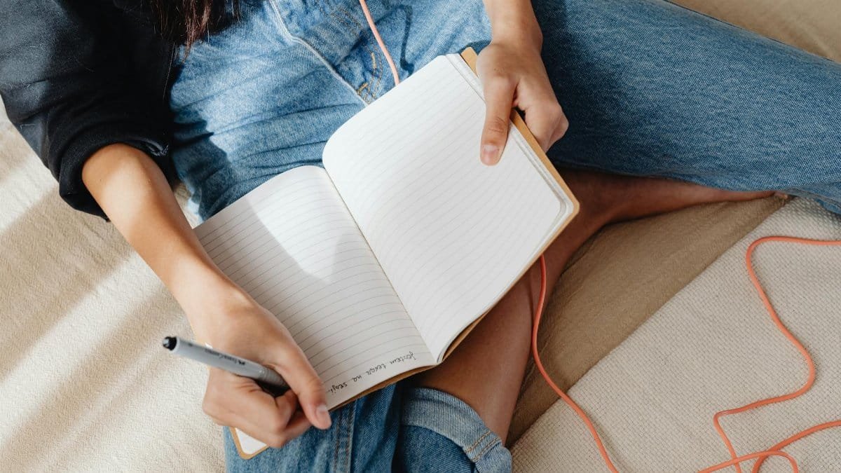 A young woman sitting comfortably, writing in a notebook while listening to music.
