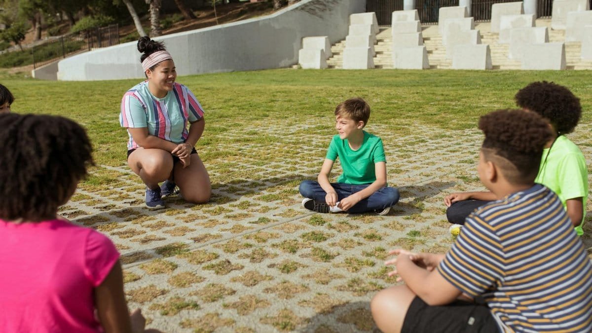 A diverse group of kids sitting in a circle outdoors, engaged in a fun summer camp activity.