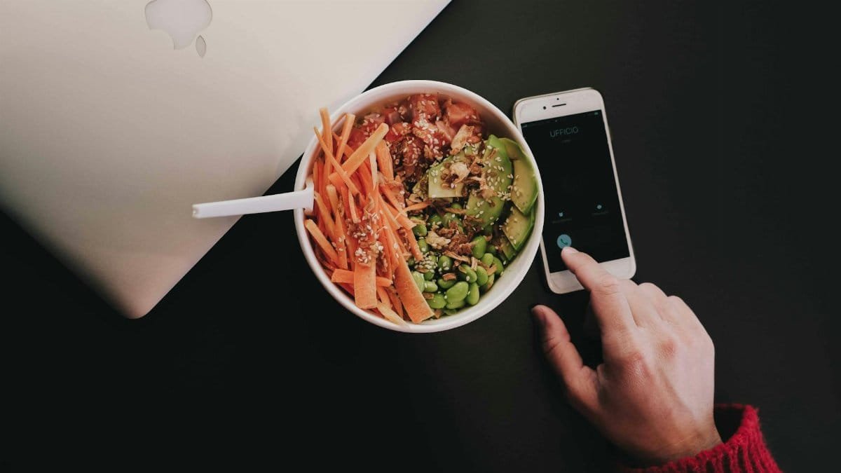A bowl of fresh salad with avocado, carrots, edamame, and quinoa on a desk beside a smartphone and laptop.