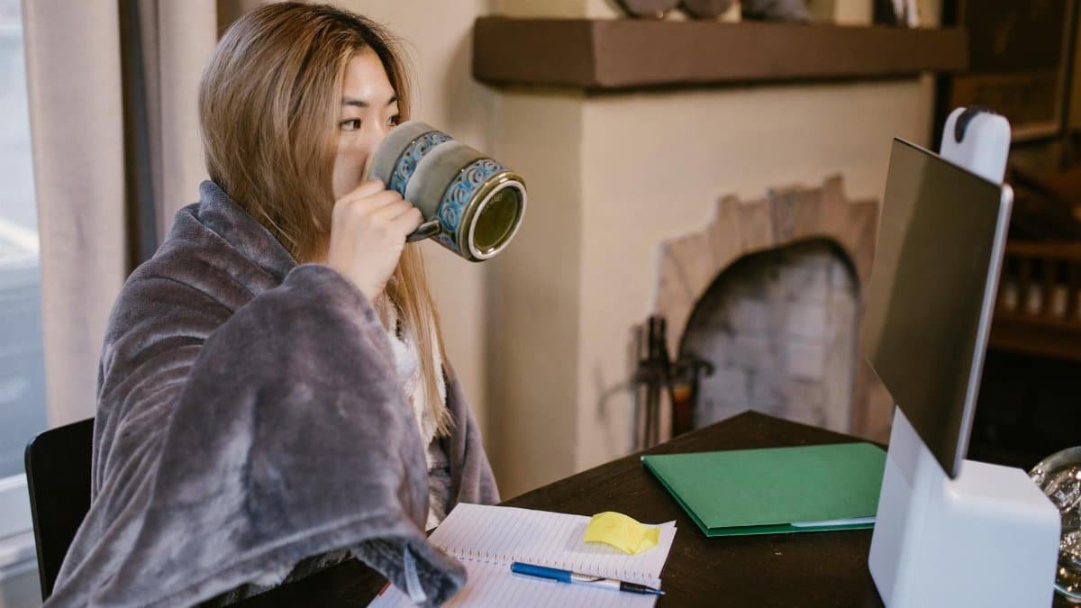 Adult woman sipping coffee while working remotely at home, with a cozy setup.
