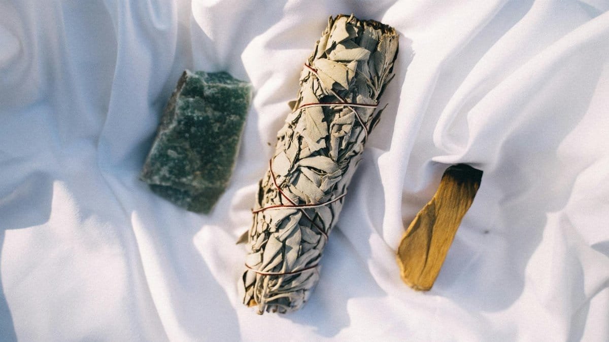 Overhead view of a calming smudging set on fabric, featuring sage, palo santo, and a healing stone.