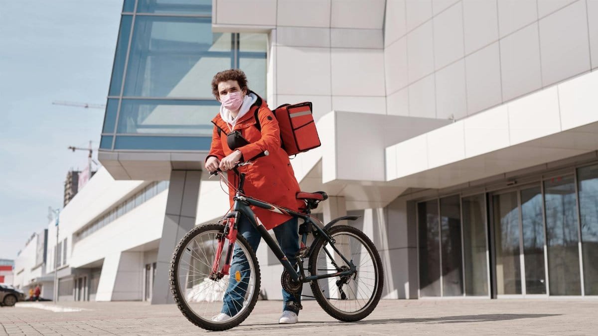 Bicycle delivery person wearing mask and thermal bag outside modern building.