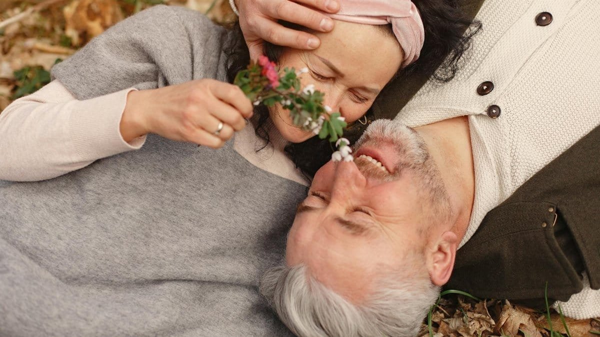 From above of cheerful senior wife wearing wide scarf and headband with flower bouquet in hand and happy elderly gray haired husband in warm clothes lying on ground with fallen leaves in park with closed eyes