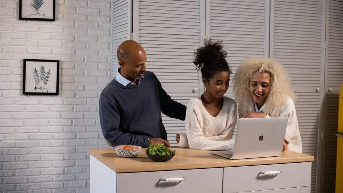 Positive African American parent and daughter surfing modern netbook while standing at wooden kitchen counter with fresh ingredients at home