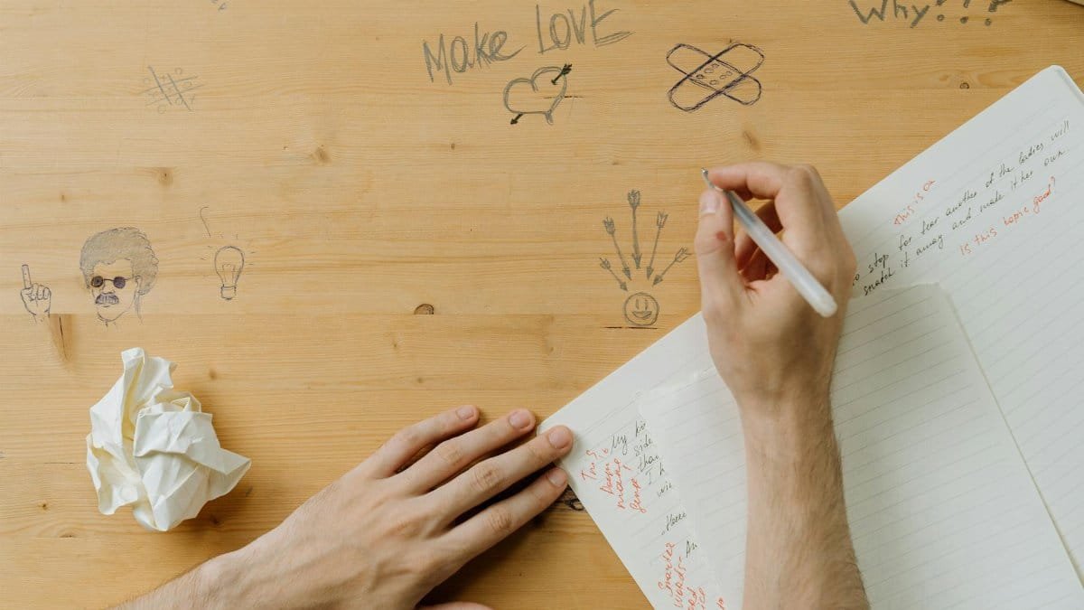 A person drawing on a wooden desk covered in creative doodles and handwritten notes.