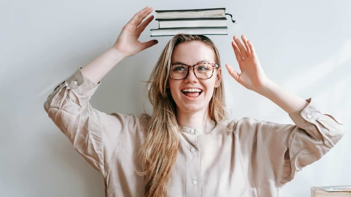 Smiling woman with glasses balancing books on her head, enjoying playful educational moments indoors.
