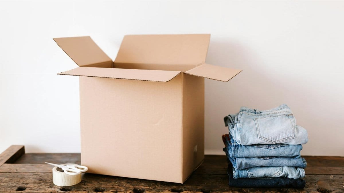 Open cardboard box on wooden table with folded jeans and packing tape nearby, ready for moving.