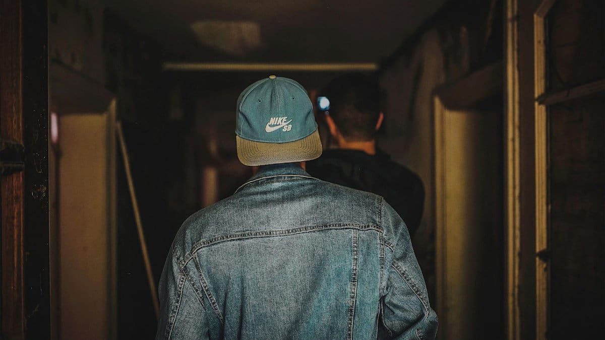 Back view of a man in a denim jacket and cap walking in a dimly lit hallway.