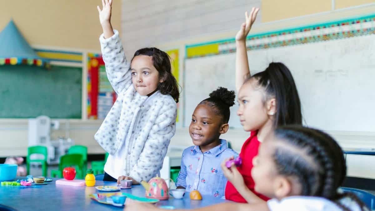Multicultural group of kids eagerly participating in a classroom setting, raising hands with smiles.