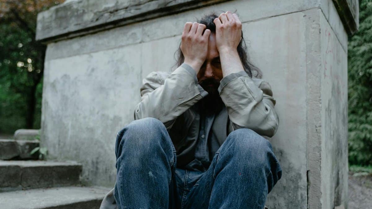 A homeless man sits in distress against a concrete wall, symbolizing poverty and isolation.