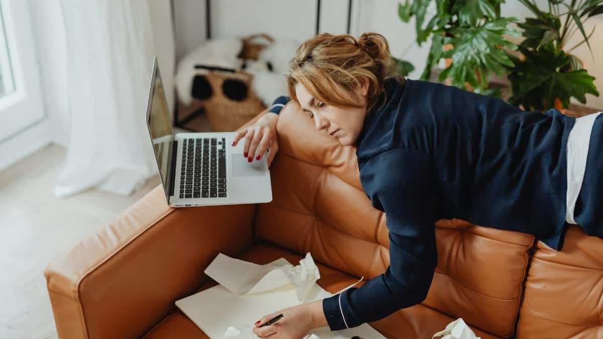 Woman resting on a couch with laptop, surrounded by papers, depicting stressed remote work lifestyle.