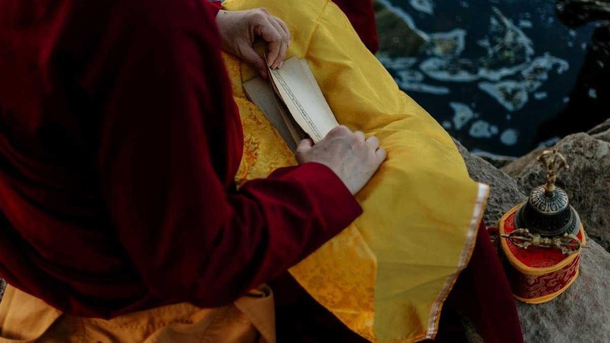 A Tibetan monk reads mantras beside a river, holding sacred objects and sitting cross-legged.