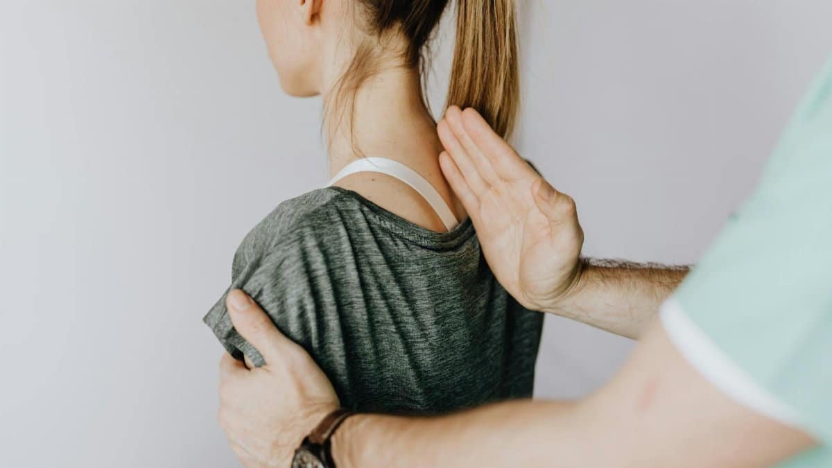 Back view of crop unrecognizable osteopath in uniform and wristwatch checking up back of slim female patient in casual wear on white background