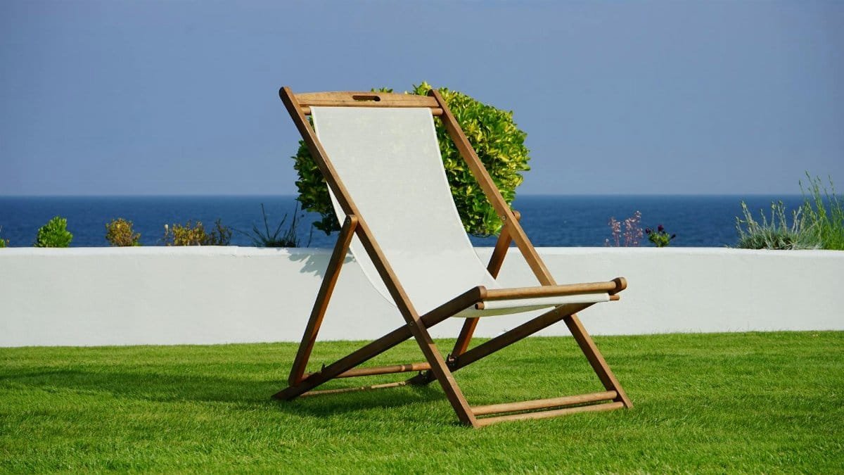 A wooden deck chair on lush green grass with a view of the blue ocean, perfect for a summer retreat.