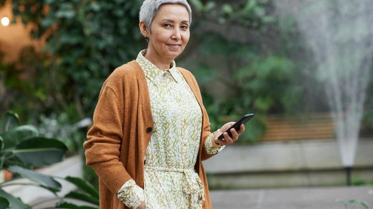 Senior woman smiling while using a smartphone in a garden setting, with greenery in the background.