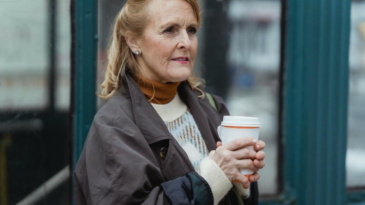 Contemplative elderly female in outerwear with hot beverage to go looking forward in town in daylight