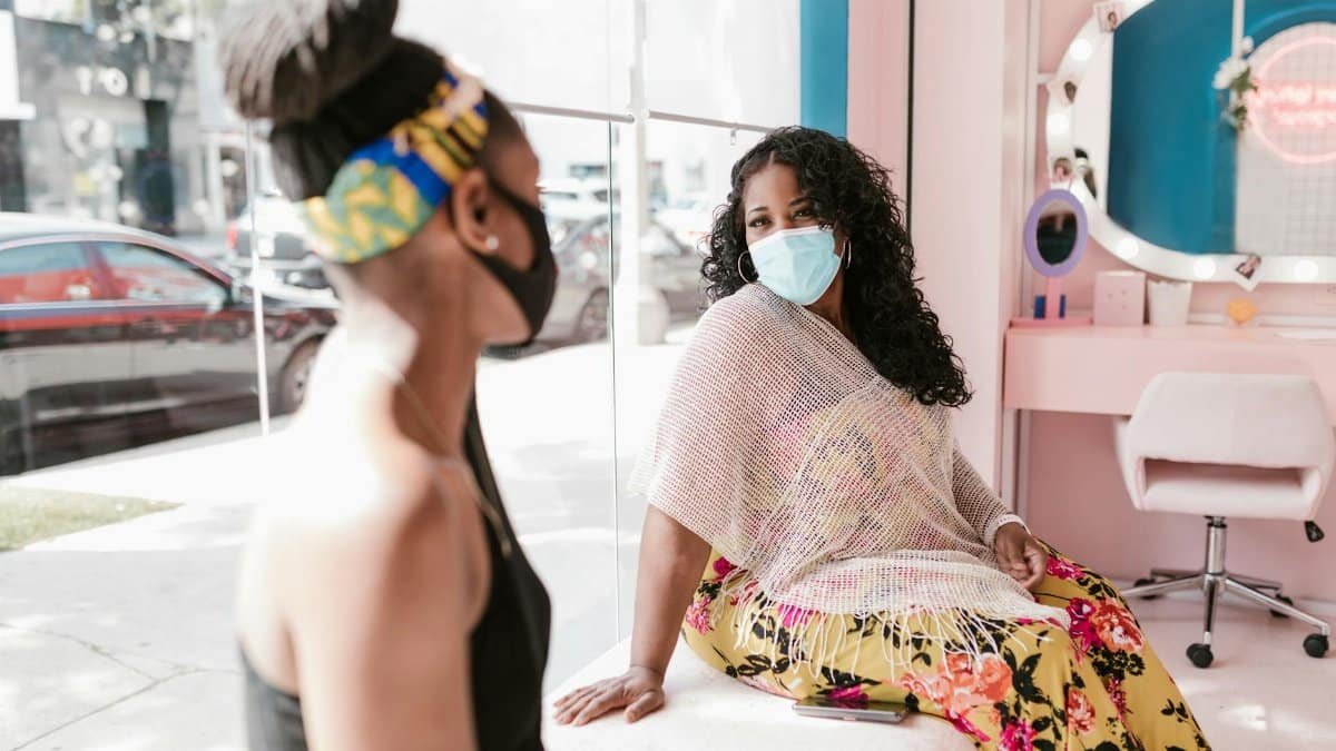 Two women wearing masks chat in a bright, stylish hair salon, embracing the new normal.